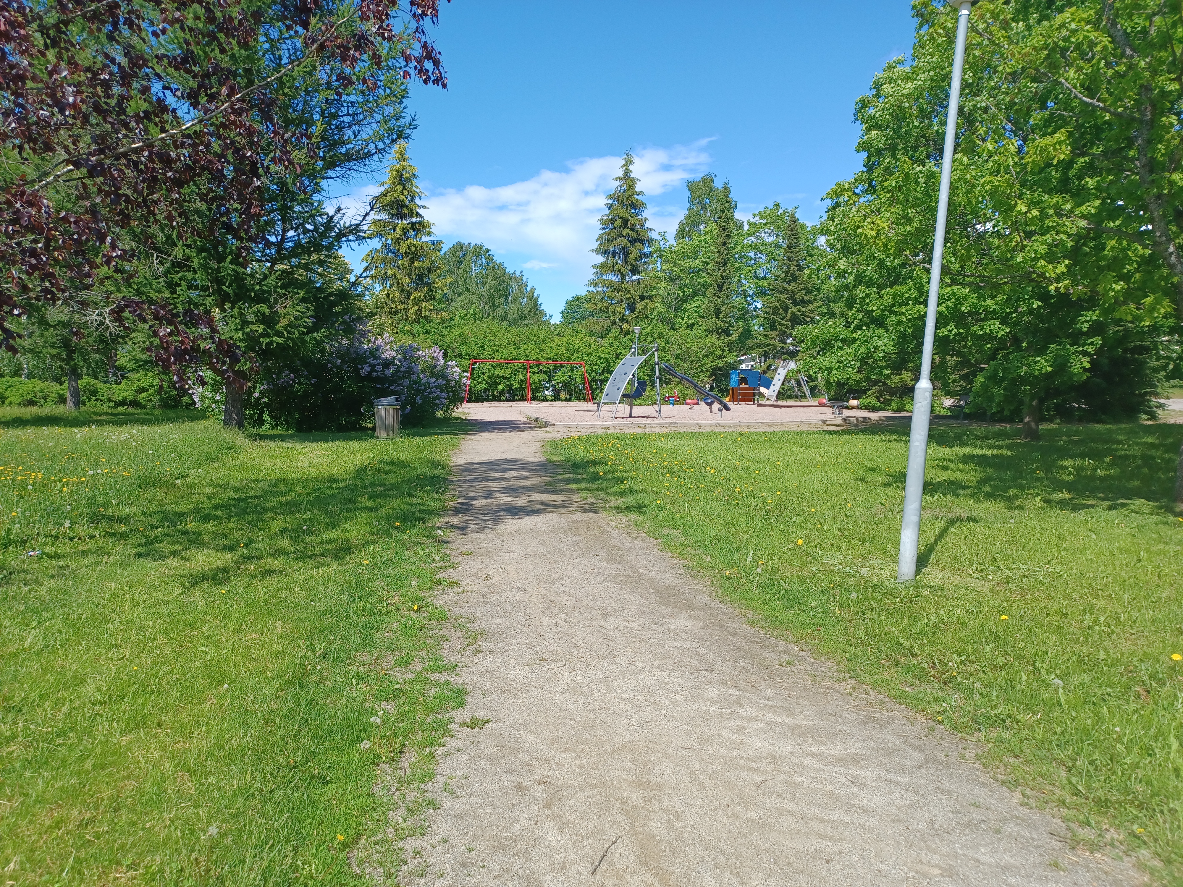 Alijuhakkalan leikkialue kesäisessä kuvassa. Ympärillä n paljon vihreää. Puistossa on keinu ja muita leikkivälineitä.