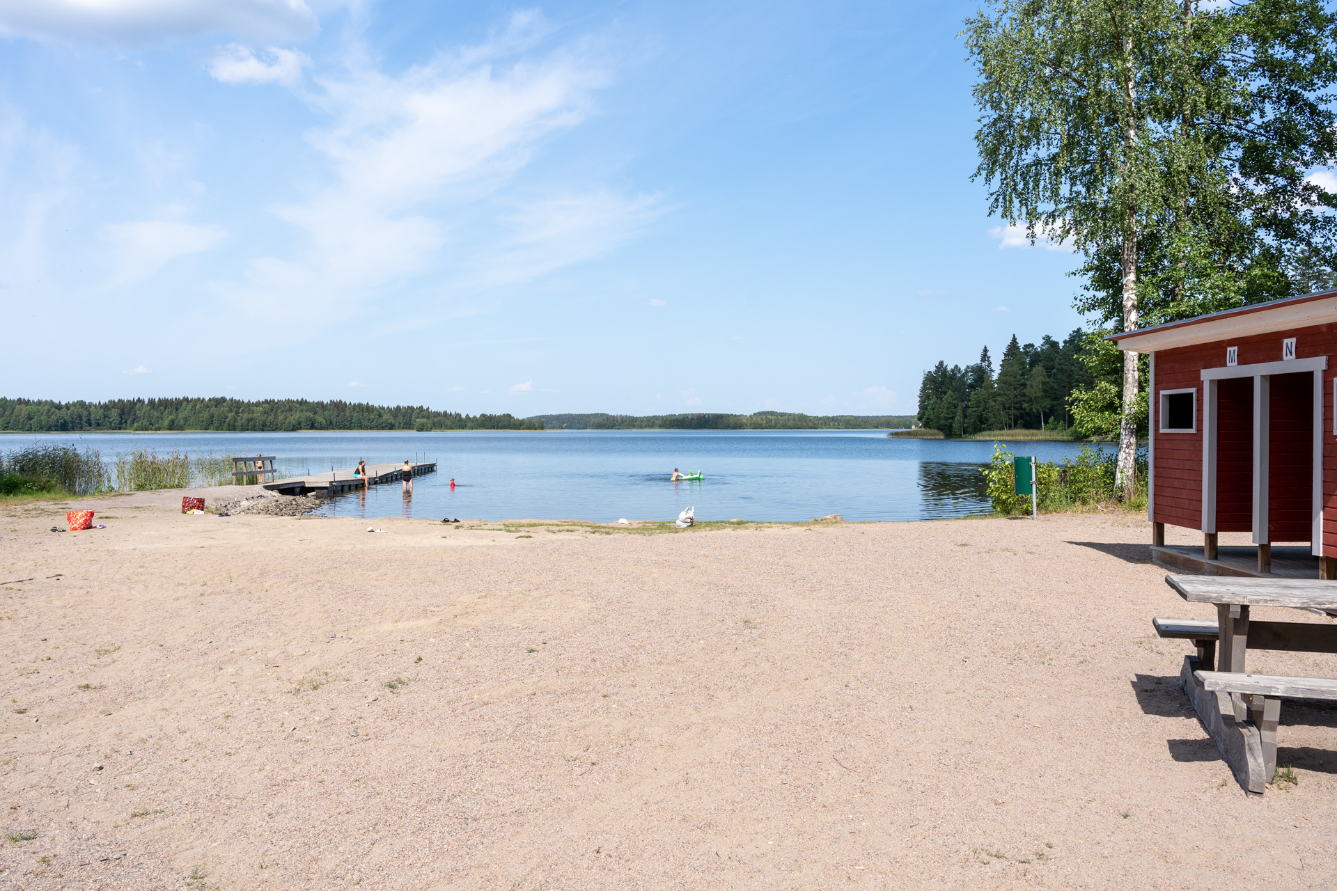 Hiekkainen uimaranta, vasemmalla näkyy pukukoppeja, oikealla laituri