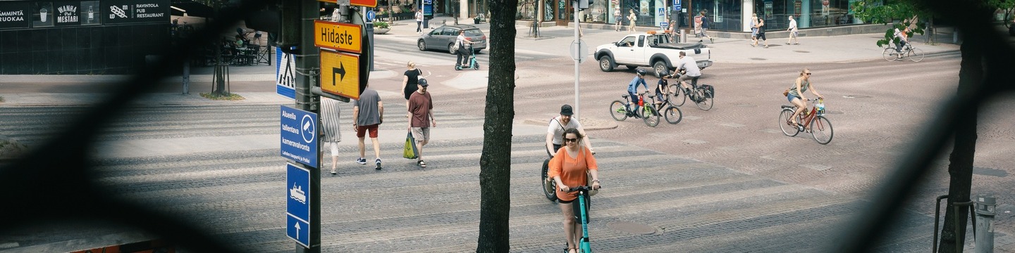 Kuvassa vilkasta kaupunkielämää Vesijärvenkadulla, jossa kuljetaan autolla, sähköpotkulaudalla, pyöräillen ja kävellen. Kuva on otettu verkkoaidan aukosta.