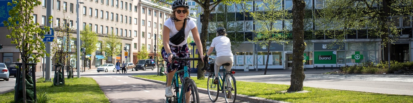 Henkilö pyöräilemässä Lahden keskustan pyörätiellä. Taustalla toinen pyöräilijä sekä muuta liikennettä. Aurinkoinen ja kaunis sää.