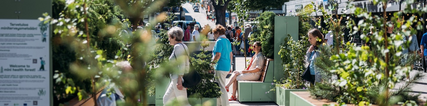 Kesäisellä Lanunaukiolla Lahdessa kävelee ihmisiä. Aukiolle on tuotu penkkejä, joihin on istutettu luonnosta tuttuja kasveja, kuten pieniä mäntyjä, jotta penkillä istujat voivat nauttia luonnosta keskellä kaupunkia